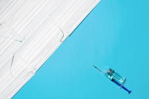 guerre conceptuelle contre le virus, vaccination de la population. flacon de vaccin, seringue et tas de masques médicaux sur fond bleu, vue de dessus photo
