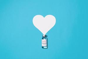 flacon de vaccin covid-19 et coeur sur fond bleu, plat. concept mondial de vaccination, de soins de santé et de médecine de la population. photo