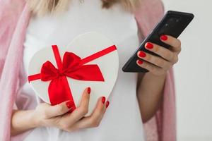 gros plan les mains de la femme avec une manucure rouge tient le coeur de la boîte-cadeau et le téléphone intelligent. service de livraison de cadeaux en ligne, achats de vacances, compliments. La Saint-Valentin photo