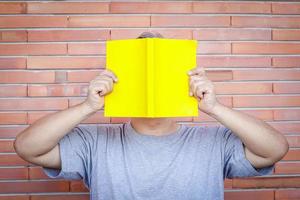 un gros homme asiatique tient un livre jaune couvrant son visage. la toile de fond est un motif de briques. concept d'éducation des adultes, développement de l'apprentissage photo