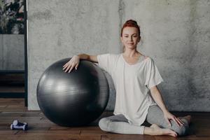 belle dame aux cheveux rouges sportive est assise avec élégance sur parquet avec fitball photo