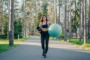 photo horizontale d'une femme mince active vêtue de vêtements de sport, porte du fitball et du karemat enroulé, marche sur la route près des arbres, va s'entraîner, respire de l'air frais, mène un mode de vie sportif sain