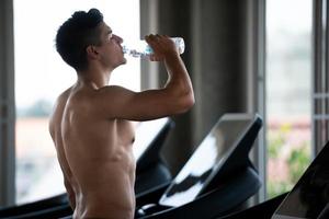 jeune homme de sport sain caucasien buvant de l'eau dans la salle de sport après l'entraînement sur tapis roulant. photo