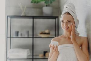 photo d'une jolie jeune femme européenne applique une crème hydratante sur le visage, a une expression heureuse, enveloppée dans une serviette de bain, se dresse sur le fond de la salle de bain à l'intérieur. concept de soins de la peau et d'hygiène