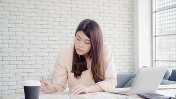 belle jeune femme asiatique souriante travaillant ordinateur portable sur le bureau dans le salon à la maison. femme d'affaires asie écrit finance de document de cahier et calculatrice au bureau à domicile profiter du temps à la maison concept. photo