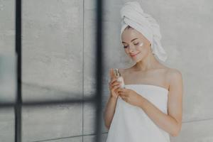 une jeune femme caucasienne satisfaite se soucie de sa beauté, applique de la crème de lotion sur le visage, se tient enveloppée dans une serviette de bain, pose dans la salle de bain de la maison contre un mur gris. soins à domicile quotidiens et concept de spa de jour photo