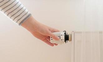 la femme régule la température à la maison avec le thermostat de chauffage pour économiser de l'énergie, gros plan avec la main. vanne de régulation de température sur radiateur. chauffage blanc au mur avec rideau dans un appartement moderne. photo