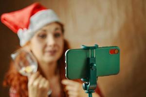 fille rousse en chapeau de père noël avec verre devant le téléphone portable. photo