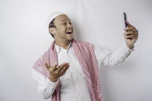 un portrait d'un homme musulman asiatique heureux souriant tout en tenant son téléphone, isolé sur fond blanc photo