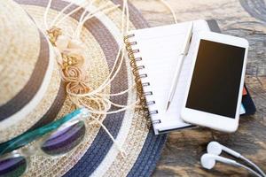 costumes d'accessoires de voyage. chapeau de plage, lunettes de soleil, bloc-notes, stylo et écouteurs, téléphone intelligent mobile avec écran vide vide sur le plan de table en bois pour préparer les voyages de vacances en été. espace pour le texte. photo