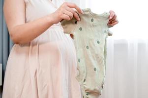 femme enceinte debout contre la fenêtre tenant des vêtements de bébé. préparer à la parentalité. photo