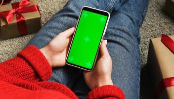 une femme méconnaissable tient un téléphone avec un écran vert entouré de cadeaux. achats de noël ou concept de médias sociaux. photo