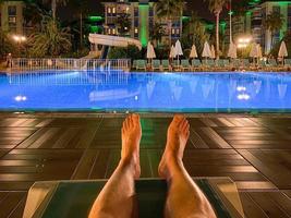 Les pieds des hommes sur une chaise longue au bord de la piscine la nuit dans un hôtel dans un pays tropical chaud de l'Est Southern Paradise Resort photo