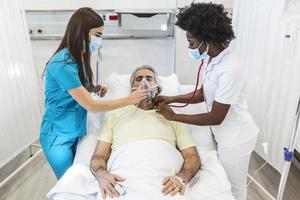 jeune médecin et infirmière portant un masque chirurgical vérifiant un patient âgé portant un masque à oxygène à pression positive pour aider à respirer dans un lit d'hôpital pendant la pandémie de covid-19 photo