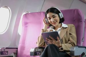 jeune femme souriante caucasienne appréciant son vol confortable tout en étant assise dans la cabine de l'avion, en écoutant de la musique dans des écouteurs et en buvant de l'eau. accès internet wifi à bord, passager près hublot photo