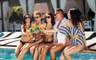 cocktails frais dans les mains. un groupe de jeunes gens heureux s'amuse dans la piscine pendant la journée photo