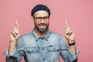 photo intérieure d'un beau mâle non rasé garde les doigts croisés, les yeux fermés, a une expression heureuse, vêtu de vêtements à la mode isolés sur fond rose. l'homme espère mieux avant un événement important