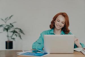 photo horizontale d'une femme indépendante au gingembre heureuse travaille sur un ordinateur portable à la maison, étudie en ligne, utilise un logiciel, surfe sur Internet, s'assoit au bureau, sourit positivement, travaille à distance