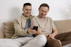 un couple gay heureux avec des vêtements décontractés passe du temps ensemble à la maison et regarde quelque chose sur un smartphone, ou passe un appel vidéo avec des amis. relations homosexuelles et amour alternatif. photo