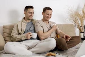 heureux couple gay avec des vêtements décontractés passant du temps ensemble à la maison, regardant un film sur l'ordinateur portable et buvant du thé. deux hommes caucasiens se détendant. relations homosexuelles et amour alternatif. photo