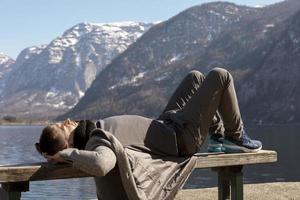 jeune homme allongé à l'extérieur sur un banc et profitant des montagnes, de la neige, du beau temps, du ciel bleu, du soleil. beau paysage. temps avec soi-même, rêve, détente, santé mentale. tourisme, vacances, voyages.