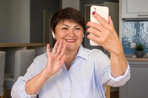 femme âgée heureuse agitant la main au téléphone comme ayant un appel vidéo avec ses amis, enfants, petits-enfants, etc. utilisation des technologies modernes photo