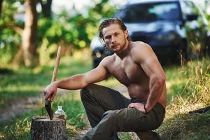 tient une hache en bois. bel homme torse nu avec un type de corps musclé est dans la forêt pendant la journée photo