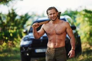 tient une hache en bois. bel homme torse nu avec un type de corps musclé est dans la forêt pendant la journée photo