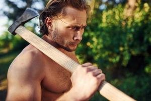 regard confiant. portrait en gros plan d'un bûcheron avec une hache à la main. bel homme torse nu avec un type de corps musclé est dans la forêt pendant la journée photo