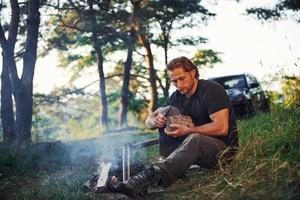 profiter d'une boisson fraîche. homme en chemise noire près du feu de camp dans la forêt pendant son week-end photo