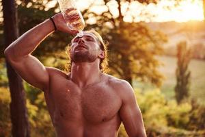 rafraîchissant avec de l'eau. bel homme torse nu avec un type de corps musclé est dans la forêt pendant la journée photo