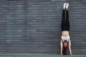 faire le poirier. jeune brune sportive avec une forme de corps mince contre un mur de briques dans la ville pendant la journée photo