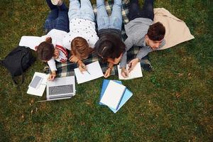 écrit sur papier. avec ordinateur portable. groupe de jeunes étudiants en vêtements décontractés sur l'herbe verte pendant la journée photo