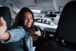 tenant la caméra. jolie fille aux cheveux noirs essayant sa toute nouvelle voiture chère dans le salon automobile photo