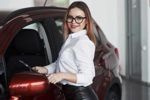 bloc-notes noir dans les mains. jolie femme en jupe noire posant près d'une voiture neuve dans un salon automobile photo