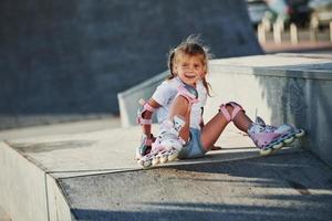 jolie petite fille avec des patins à roulettes à l'extérieur est assise sur la rampe pour les sports extrêmes photo