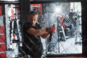 vue de face. le jeune boxeur en bandages rouges fait de l'exercice. dans le gymnase sur la cage photo