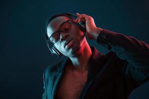 profiter de l'écoute de la musique dans les écouteurs. dans des verres. éclairage néon futuriste. jeune homme afro-américain dans le studio photo