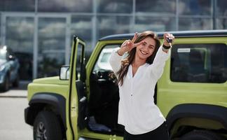 expression positive du visage. détient les clés de son nouveau véhicule. une jeune femme en tenue officielle blanche se tient devant une automobile verte à l'extérieur photo