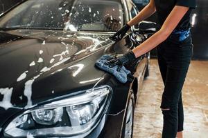 vue rapprochée du véhicule en train de se faire essuyer. une automobile noire moderne est nettoyée par une femme à l'intérieur d'une station de lavage de voiture photo