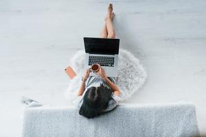 vue de dessus d'une jeune femme en vêtements décontractés assise seule à la maison avec un ordinateur portable et du thé photo