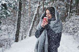 grosse écharpe chaude sur la tête. fille en vêtements chauds avec une tasse de café se promener dans la forêt d'hiver photo