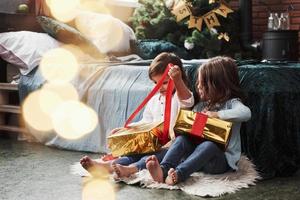 enfin je peux l'ouvrir. vacances de noël avec des cadeaux pour ces deux enfants assis à l'intérieur dans la belle chambre près du lit photo