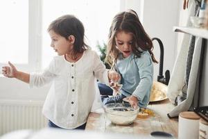 mélanger la pâte et détient des instruments colorés spéciaux. amis d'âge préscolaire apprenant à cuisiner avec de la farine dans la cuisine blanche photo