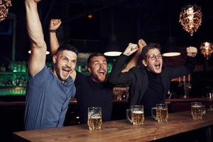 célébrant la victoire. trois fans de sport dans un bar regardent le football. avec de la bière dans les mains photo