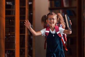 courir en avant. jolie petite fille avec sac à dos se tient dans la bibliothèque pleine de livres. conception de l'éducation photo