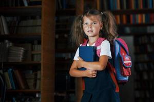 jolie petite fille avec sac à dos se tient dans la bibliothèque pleine de livres. conception de l'éducation photo