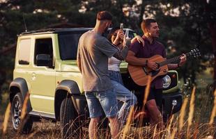 jouer de la guitare acoustique et chanter. un groupe d'amis joyeux passe un bon week-end à une journée ensoleillée près de leur voiture verte à l'extérieur photo