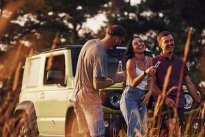 parler et tenir des boissons dans les mains. un groupe d'amis joyeux passe un bon week-end à une journée ensoleillée près de leur voiture verte à l'extérieur photo