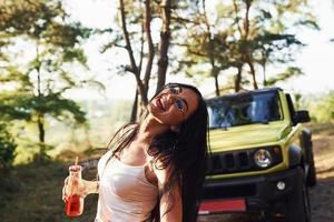 posant pour la caméra. beaux cheveux longs. blonde avec de l'alcool à la main se promener dans la forêt contre la jeep verte photo